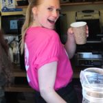 Photo of a woman in pink tshirt holding a coffee cup next to coffee making equipment