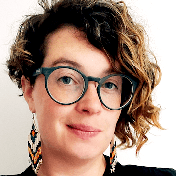 Photo of Cathy, who is white with asymmetrical wavy hair, wearing large round glasses and earrings