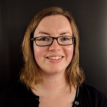 Head photo of Hannah Ross, who is smiling at the camera. The background is very dark. She has light brown hair and glasses.